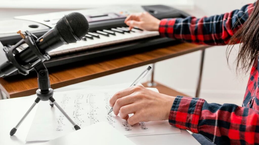 Close-up of a hand writing notes, resembling the process of composing a song. A microphone is visible, with a blurred piano in the background