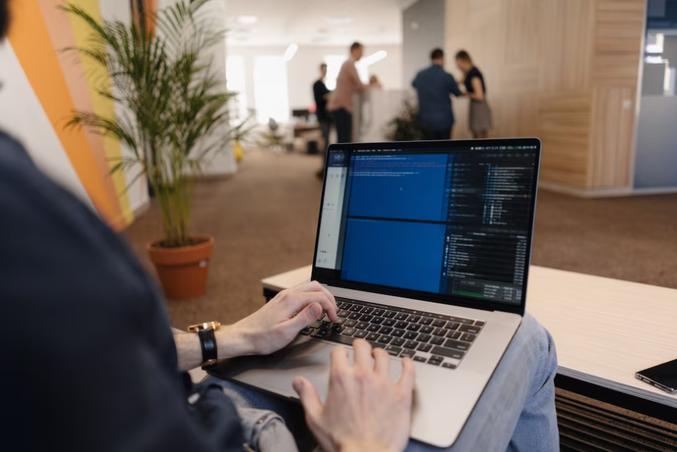 man sitting on the sofa and typing on the laptop with code on it, people in the office behind